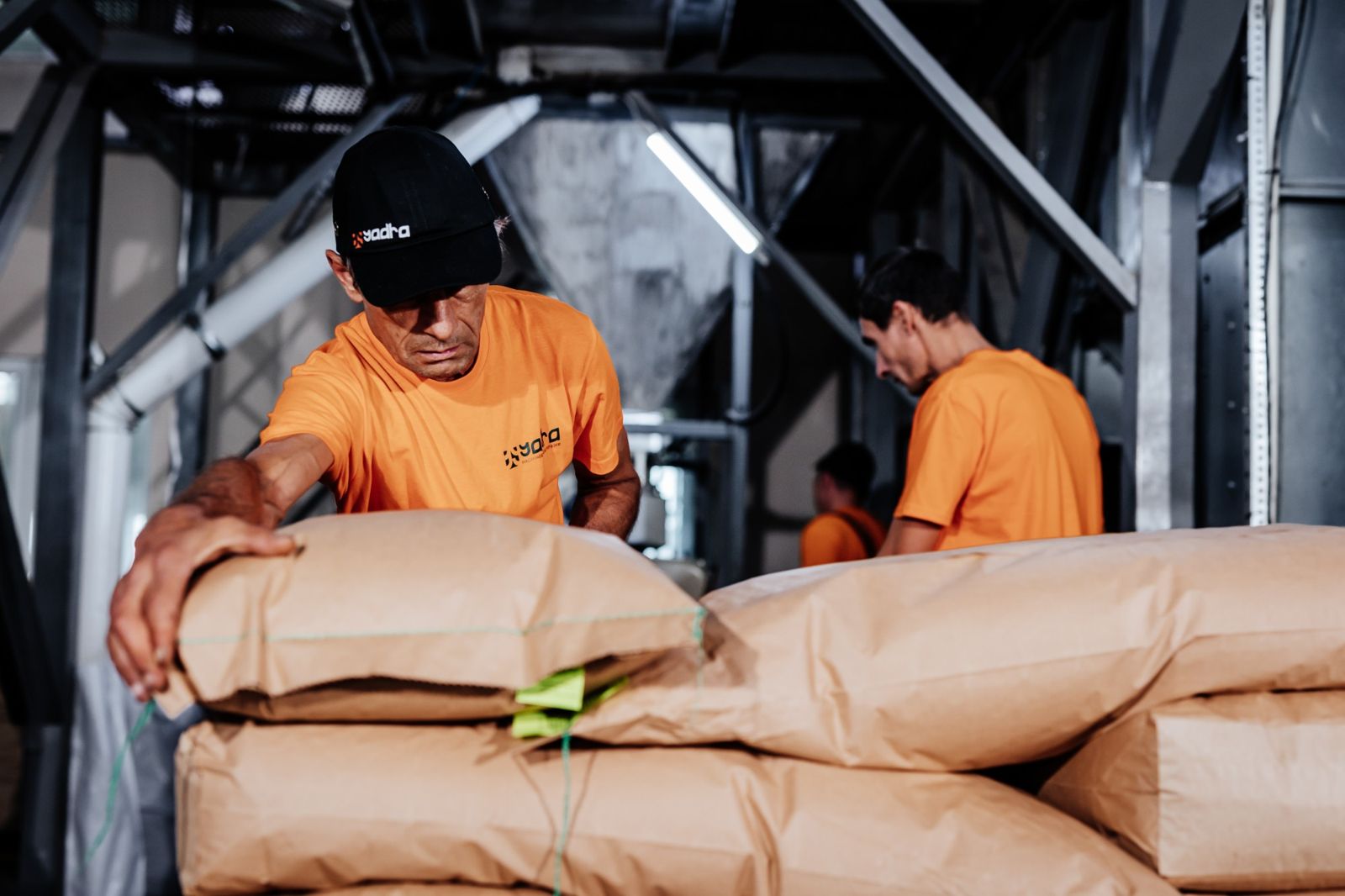 Two Yadra workers in orange shirts stacking paper sacks in the processing plant